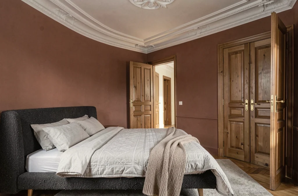 Chambre haussmannienne avec rosace en plâtre préservée, portes anciennes dorées et mobilier contemporain en bouclé charbon sur fond terracotta